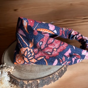 May include: A blue headband with a floral pattern in shades of orange, pink, and white. The headband is made of a soft, stretchy fabric and has a knotted design.