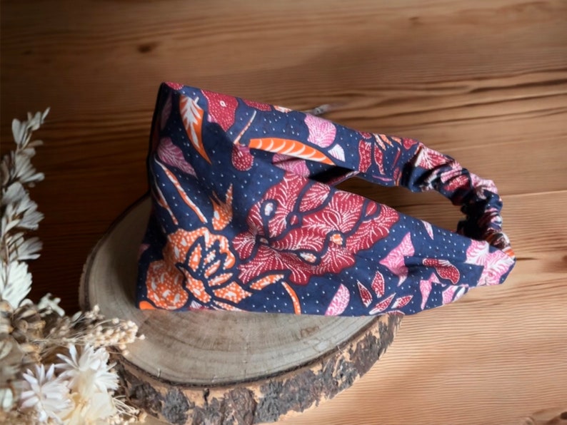 May include: A blue headband with a floral pattern in shades of orange, pink, and white. The headband is made of a soft, stretchy fabric and has a knotted design.
