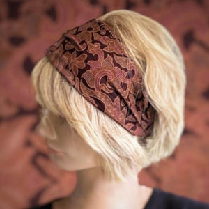 Op de afbeelding: Een vrouw met kort blond haar draagt een bruin en gouden geprint haarband. Het haarband is gemaakt van stof en heeft een paisley-ontwerp.