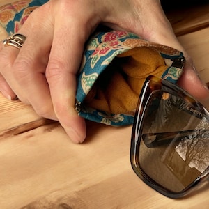 May include: A teal and floral patterned fabric eyeglass case with a gold lining. The case is open and a pair of brown tortoise shell sunglasses with brown lenses are partially inside. The sunglasses have the text "E.WESTV" and "400 CE CAT.3" printed on the frame.