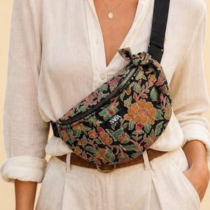 Puede incluir: Un bolso de cintura negro con estampado floral, cremallera y correa negras. La bolsa tiene un estampado floral en tonos naranja, rosa y verde. La palabra "SABA" está impresa en la parte delantera de la bolsa.