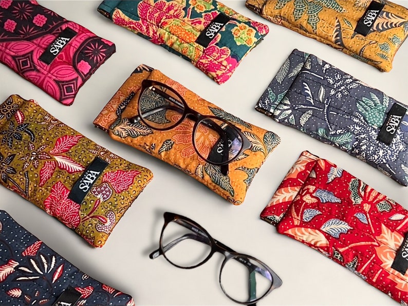 May include: Assortment of colorful patterned eyeglass cases with the word "SABA" printed on each case.  The cases are made of fabric and have a variety of floral and geometric designs.  Two pairs of eyeglasses are also shown in the image.