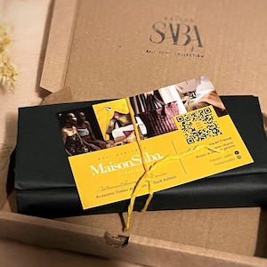 May include: An open cardboard box containing a wrapped package and a yellow card. The card features the text "Maison Saba" and images of bags. The package is wrapped in black paper and tied with a yellow string.