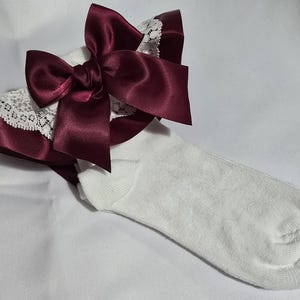 Può includere: Calzini alla caviglia bianchi con un fiocco di raso bordeaux e rifiniture in pizzo. Il fiocco è grande e centrato, aggiungendo un tocco decorativo ai calzini. Le rifiniture in pizzo sono bianche.