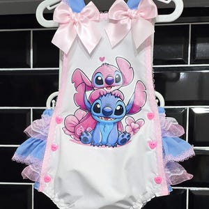 May include: A white baby onesie with a blue and pink Stitch character print. The onesie has a blue and pink ruffled trim and pink satin bows on the straps.
