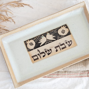 Puede incluir: Una bandeja rectangular de cerámica blanca con un diseño en blanco y negro de granadas y pájaros. La bandeja tiene las palabras hebreas "L'Shanah Tovah" escritas en ella.