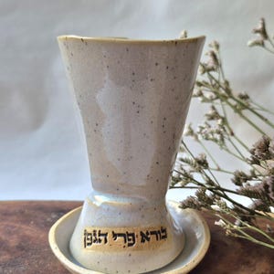 Pode incluir: Uma taça de Kiddush de cerâmica branca com um pires correspondente. A taça e o pires têm um esmalte salpicado e o pires tem as palavras hebraicas "ברוך אתה" escritas nele.