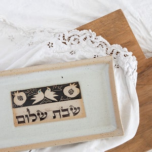 Puede incluir: Una bandeja rectangular de servir de cerámica blanca con un diseño en blanco y negro de granadas y pájaros. La bandeja tiene un borde beige y las palabras "Shalom L'ch" están escritas en hebreo en la bandeja.