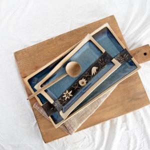 May include: Two blue ceramic rectangular serving trays with black and gold floral designs, stacked on a wooden cutting board with a wooden spoon.