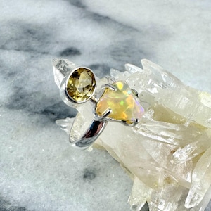 Könnte beinhalten: Ein silberner Ring mit einem hellgelben und weißen Opal-Edelstein. Der Ring liegt auf einem Bett aus weißen Kristallen.