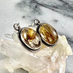 May include: A pair of silver earrings with oval-shaped gold rutilated quartz stones. The stones have a clear quartz base with gold inclusions.