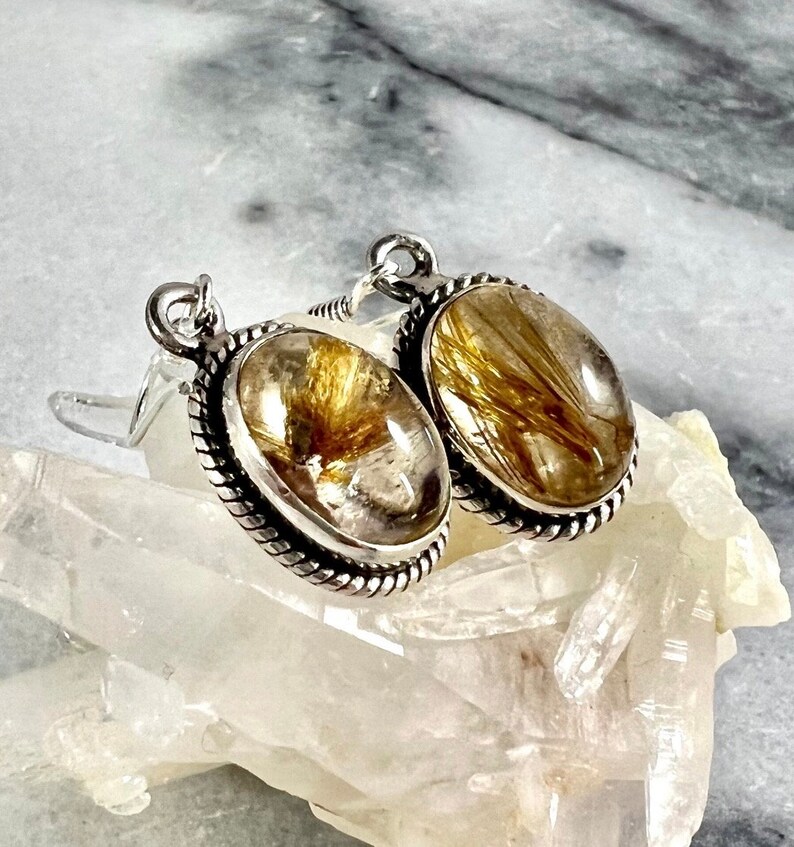 May include: A pair of silver earrings with oval-shaped gold rutilated quartz stones. The stones have a clear quartz base with gold inclusions.