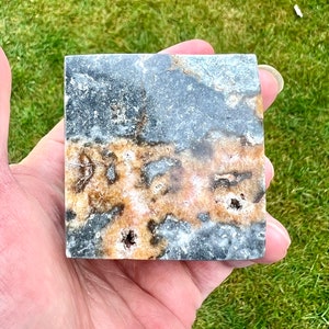May include: A square piece of polished black and brown stone with white and orange veins. The stone has a rough texture and is about 5 cm wide.