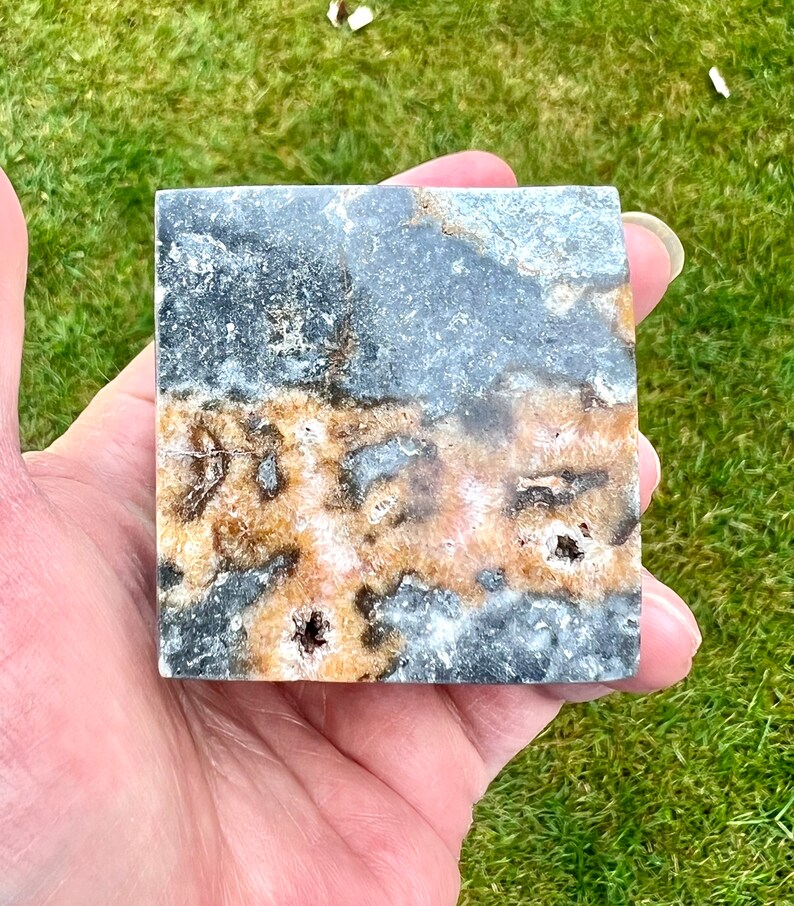May include: A square piece of polished black and brown stone with white and orange veins. The stone has a rough texture and is about 5 cm wide.