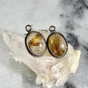 May include: A pair of silver earrings with oval-shaped clear quartz stones containing gold rutile inclusions. The earrings have a rope-like border around the stones.
