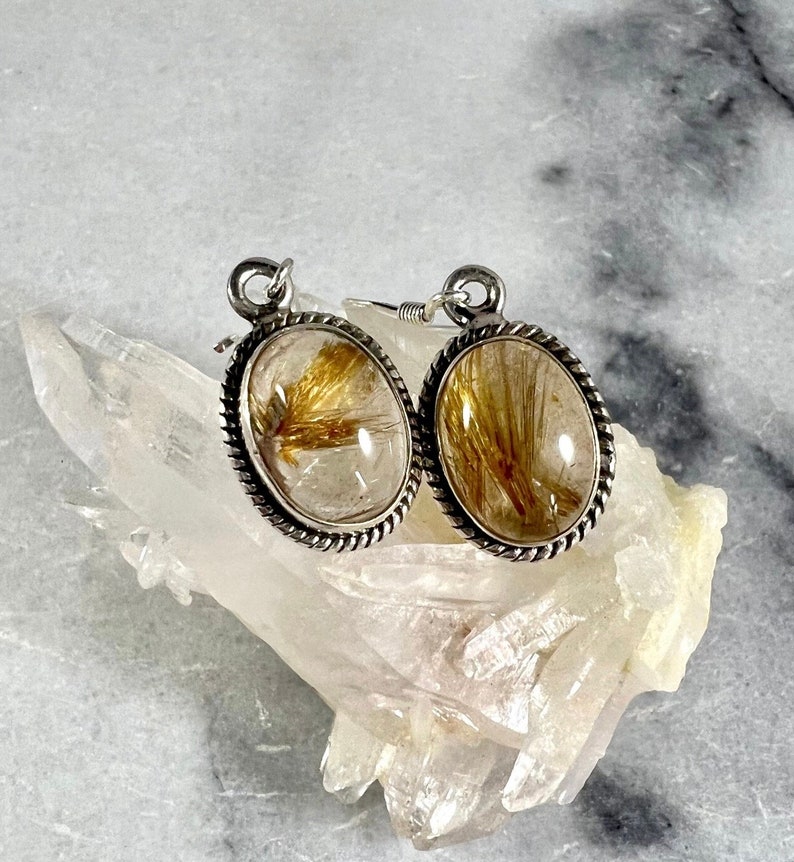 May include: A pair of silver earrings with oval-shaped clear quartz stones containing gold rutile inclusions. The earrings have a rope-like border around the stones.