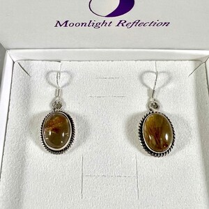 May include: A pair of silver earrings with oval-shaped brown and gold stones set in a silver bezel. The earrings are in a white box with the text "Moonlight Reflection" on the lid.