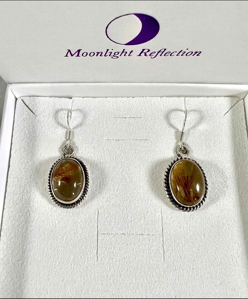 May include: A pair of silver earrings with oval-shaped brown and gold stones set in a silver bezel. The earrings are in a white box with the text "Moonlight Reflection" on the lid.