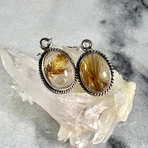 May include: A pair of silver earrings with oval-shaped clear quartz stones containing gold rutile inclusions. The earrings have a rope-like border around the stones.