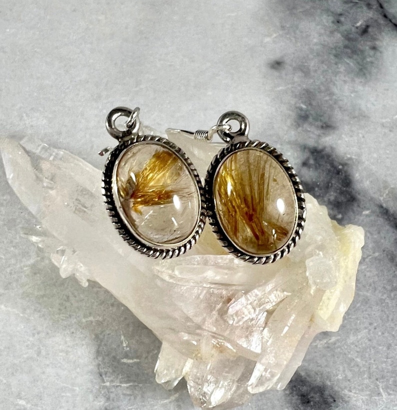 May include: A pair of silver earrings with oval-shaped clear quartz stones containing gold rutile inclusions. The earrings have a rope-like border around the stones.