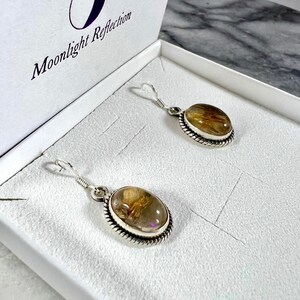 May include: A pair of silver earrings with oval-shaped, clear quartz stones with gold inclusions. The earrings are in a white box with the text "Moonlight Reflection" on the lid.