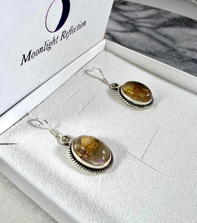 May include: A pair of silver earrings with oval-shaped, clear quartz stones with gold inclusions. The earrings are in a white box with the text "Moonlight Reflection" on the lid.