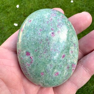 May include: A green and pink tumbled stone with a smooth, polished surface. The stone is speckled with small, pink inclusions.