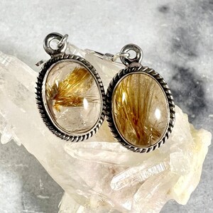 May include: Silver earrings with a clear quartz stone containing gold rutile inclusions. The earrings have a rope-like design around the edge of the stone.