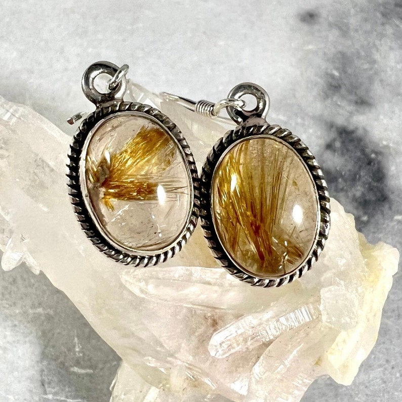 May include: Silver earrings with a clear quartz stone containing gold rutile inclusions. The earrings have a rope-like design around the edge of the stone.