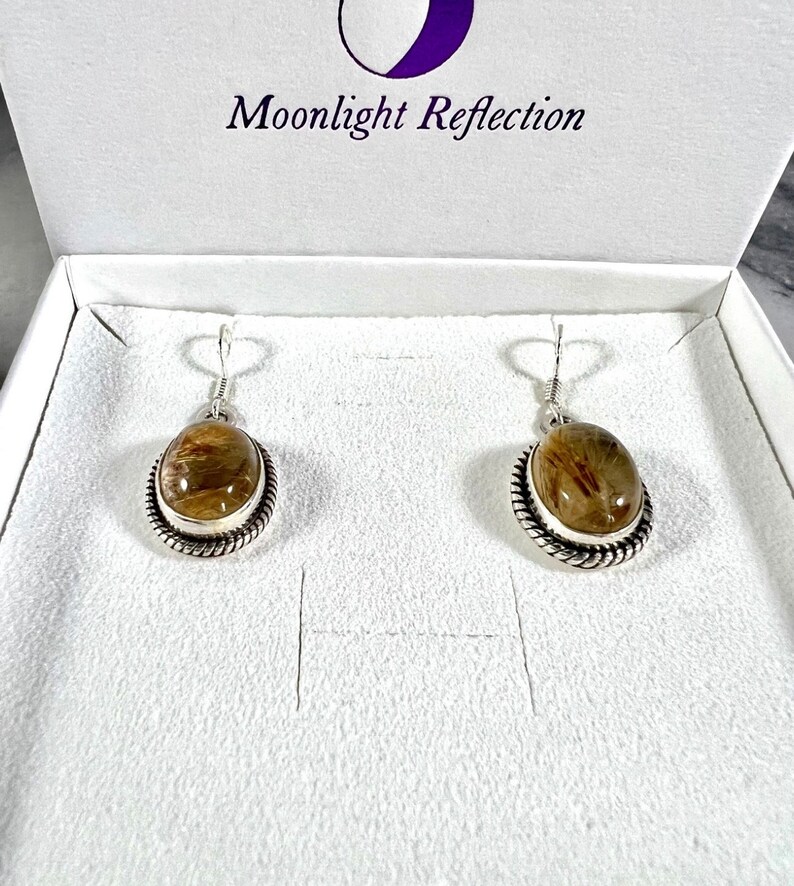 May include: A pair of silver earrings with brown and clear quartz stones. The earrings are in a white box with the text "Moonlight Reflection" on the lid.