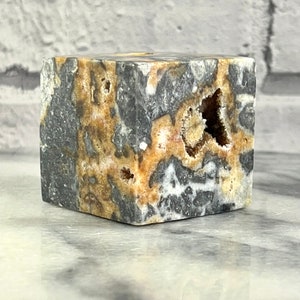 May include: A cube-shaped, polished stone with a grey, white, and brown marbled pattern. The stone has several holes and cavities, revealing a white crystalline structure.