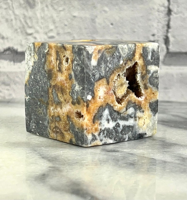 May include: A cube-shaped, polished stone with a grey, white, and brown marbled pattern. The stone has several holes and cavities, revealing a white crystalline structure.