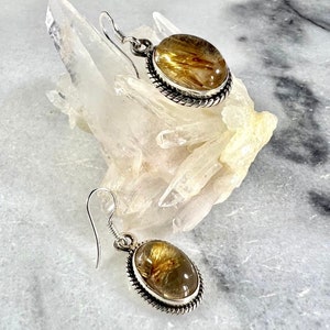May include: A pair of silver earrings with oval-shaped golden rutilated quartz stones. The stones have a clear, glassy appearance with golden inclusions.