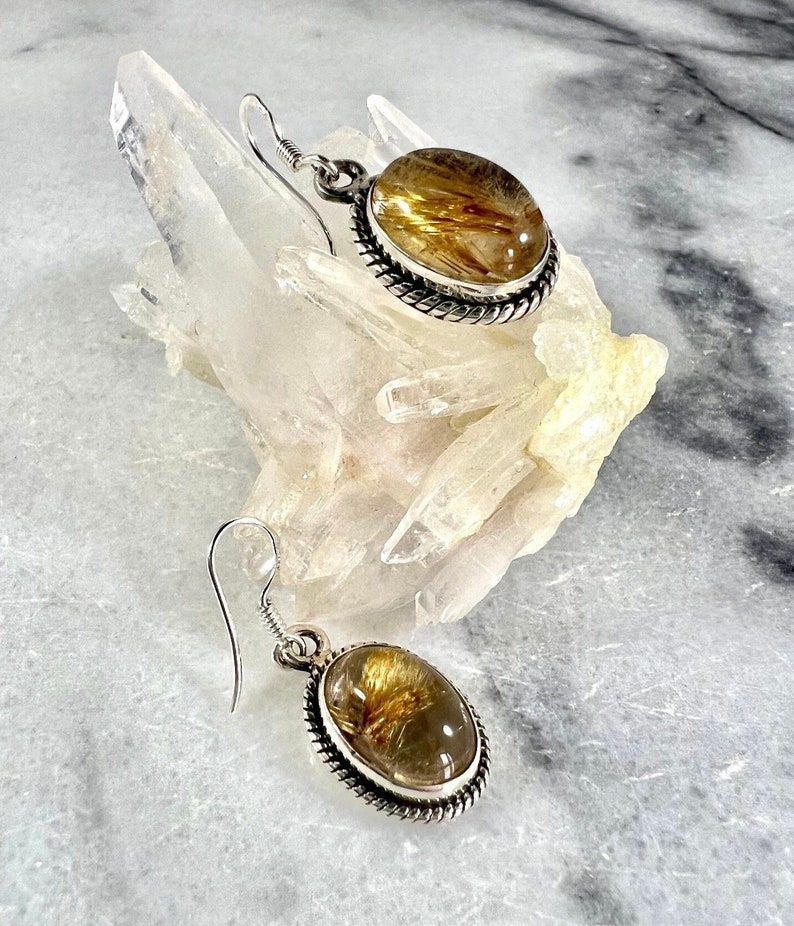 May include: A pair of silver earrings with oval-shaped golden rutilated quartz stones. The stones have a clear, glassy appearance with golden inclusions.