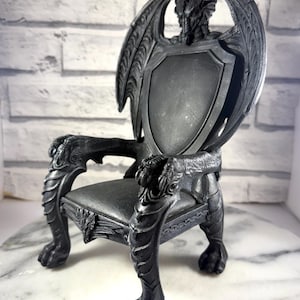 May include: A black fantasy throne with dragon-inspired details. The chair features a dragon head at the top, wings forming the back, and clawed feet. The seat is a dark grey colour, and the chair is made of a solid material.