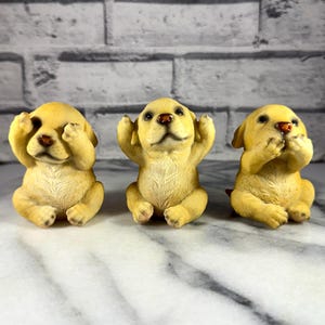 May include: Three yellow Labrador puppy figurines in the 'See No Evil, Hear No Evil, Speak No Evil' pose. Each puppy is sitting with its paws covering its eyes, ears, and mouth. The figurines have brown noses and are on a white surface.