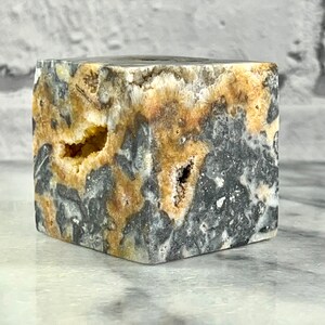 May include: A small, cube-shaped, grey and yellow geode with a rough, natural texture. The geode has several small holes and cavities.