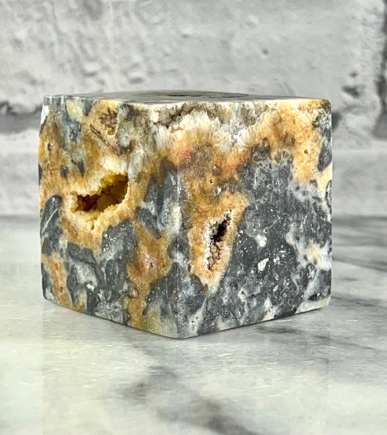 May include: A small, cube-shaped, grey and yellow geode with a rough, natural texture. The geode has several small holes and cavities.