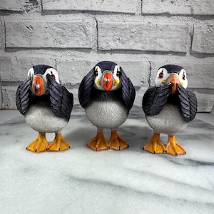 May include: Three black and white puffin figurines, each with one hand covering their eyes, one hand covering their ears, and one hand covering their mouth. The figurines are arranged in a row on a white marble surface.