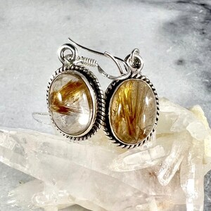 May include: Silver earrings with oval-shaped clear quartz stones containing gold rutile inclusions. The earrings have a rope-like design around the edges of the stones.