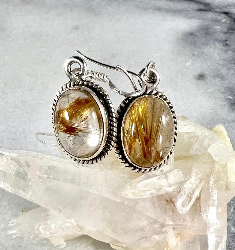 May include: Silver earrings with oval-shaped clear quartz stones containing gold rutile inclusions. The earrings have a rope-like design around the edges of the stones.