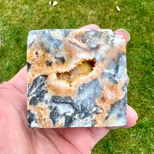 May include: A square-shaped agate geode with a natural opening revealing a crystal cave. The geode has a brown, white, and black colour pattern.