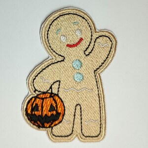 May include: An embroidered patch of a gingerbread man holding a jack-o'-lantern. The gingerbread man is smiling and has white icing eyes and a red icing mouth. The jack-o'-lantern is orange with a black face.