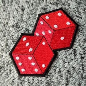 May include: Two red dice with white dots, sewn onto a gray fabric background. The dice are stacked on top of each other.