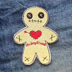 May include: A yellow voodoo doll patch with black cross eyes and mouth stitches. A red heart is pierced with a pin. The words "Ex-boyfriend" are embroidered in red below the heart. The patch is on a blue denim background.