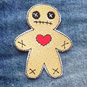 May include: Embroidered voodoo doll patch with a tan body, black stitched eyes and mouth, and a red heart. The patch has a white border and black stitched details on the arms and legs. The patch is on a blue denim background.