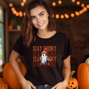 May include: A black t-shirt with the text "STAY SPOOKY" stacked vertically, featuring a cartoon ghost wearing a knit cap and holding a pumpkin. The shirt is worn by a person in front of several orange pumpkins.