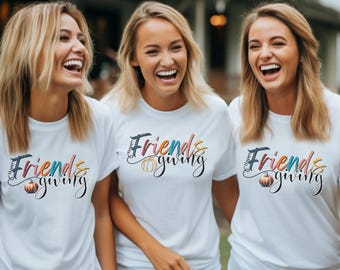 May include: Three women wearing white t-shirts with the text "Happy Friendsgiving" in a colorful, stylized font. Each shirt features a pumpkin graphic. The women are smiling and laughing, suggesting a joyful gathering.