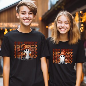 May include: Black t-shirts with a Halloween-themed graphic. The design features a cartoon ghost wearing a knit cap and holding a pumpkin and a drink, with the words "Stay Spooky" in orange and brown.
