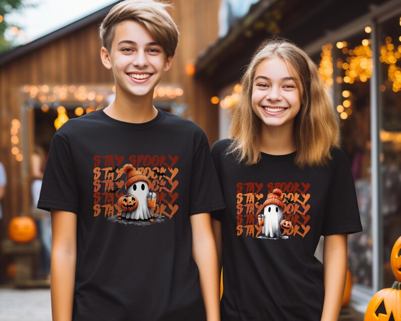 May include: Black t-shirts with a Halloween-themed graphic. The design features a cartoon ghost wearing a knit cap and holding a pumpkin and a drink, with the words "Stay Spooky" in orange and brown.